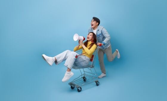 Young woman sitting in shopping trolley with a megaphone. She is being pushed by a man and they are both surprised and happy.